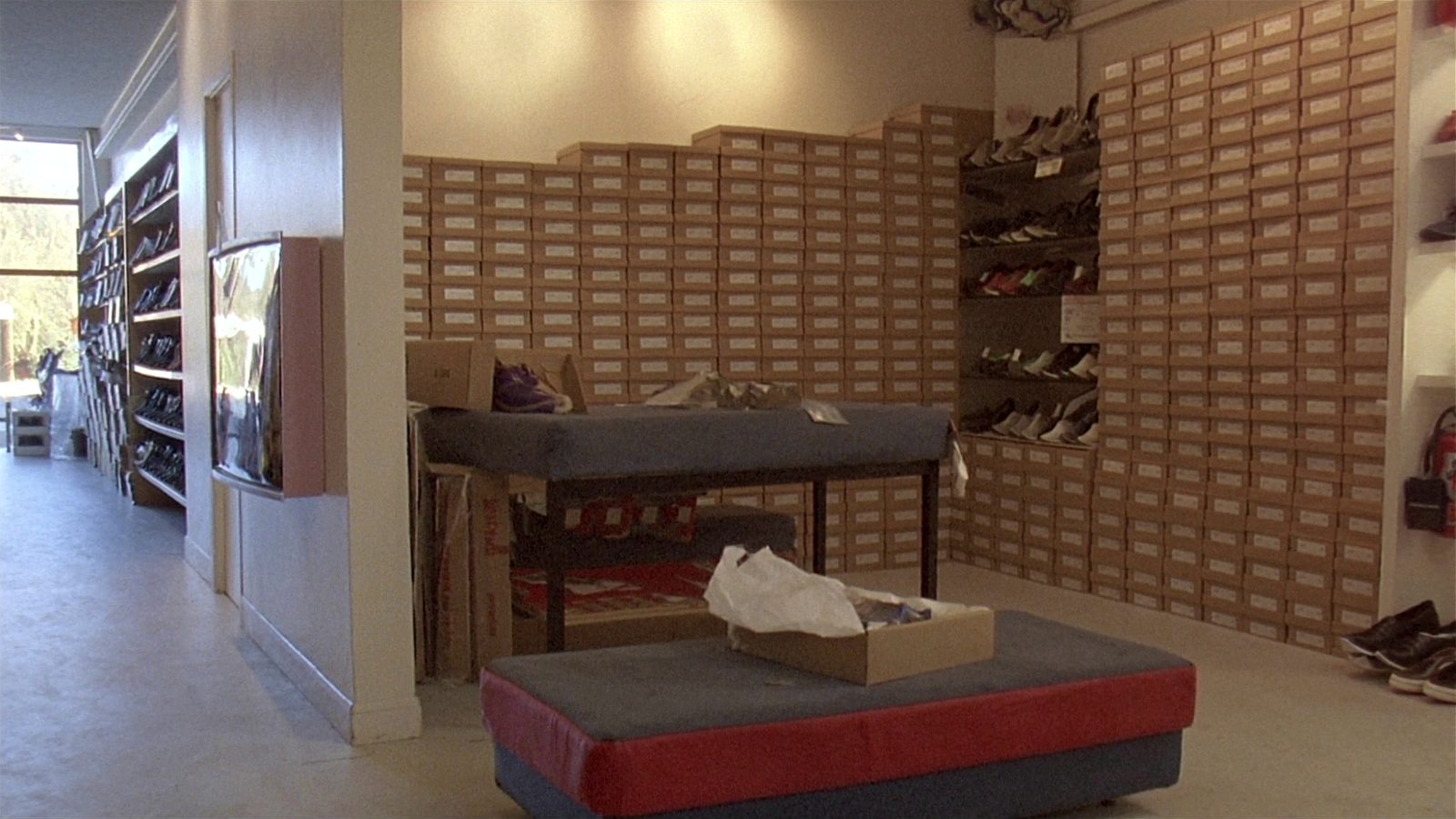 Inside a shoe shop: two low padded fitting benches on a pale floor, one holding an open cardboard box with tissue paper in it, and behind them walls of plain kraft shoe boxes stacked from floor to ceiling, with daylight coming in from the shopfront at the left.