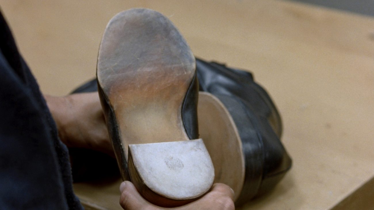 A hand holding a worn men's leather dress shoe sole-upward on a pale counter, with the second shoe lying behind it. The forepart of the sole is scuffed grey and the heel is a paler replacement patch.